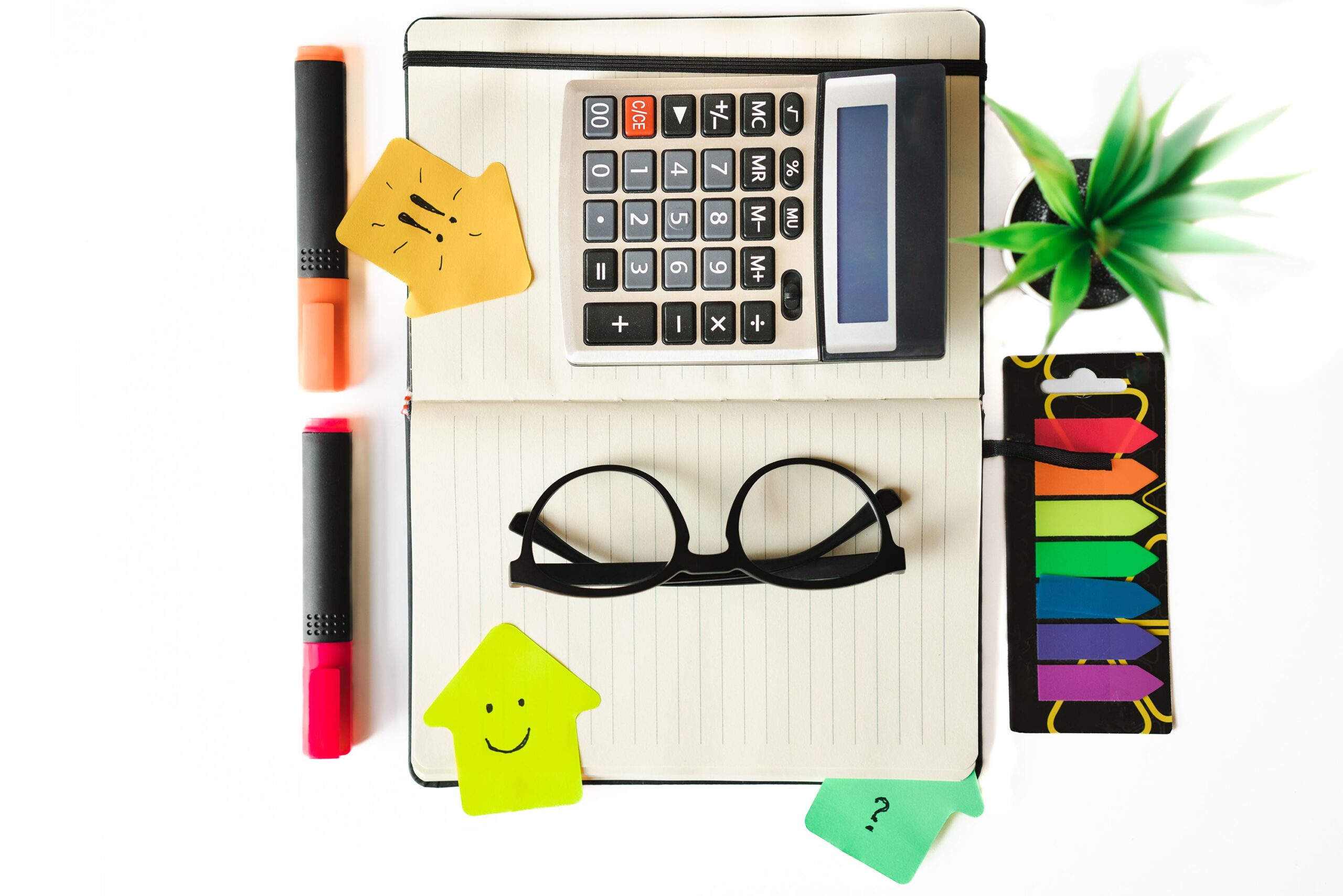 Top view of a creative workspace featuring a calculator, notebook, glasses, and colorful stationery on a white background.